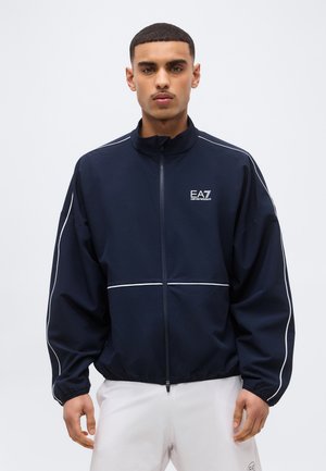 Νεαρός άνδρας που φοράει μπλε σκούρο μπουφάν EA7 με φερμουάρ και λευκές ρίγες, καθώς και λευκό παντελόνι, στέκεται μπροστά σε ένα απλό ανοιχτό φόντο.