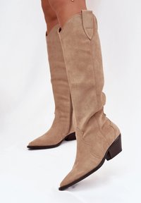 Bottes en daim beige, montantes jusqu'au genou, avec des bouts pointus, de petits talons carrés et des coutures décoratives sur la tige. Présente un design légèrement ample.