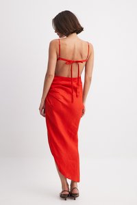 Abito rosso in lino con design a schiena bassa e spalline regolabili. Presenta uno spacco laterale e una silhouette fluida. Indossato con sandali con tacco e lacci.