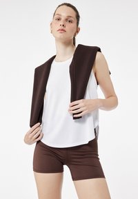Hvid ærmeløs top med rund halsudskæring, parret med brune tætsiddende shorts. Brunt stof hænger over skuldrene og skaber et lagdelt udseende.