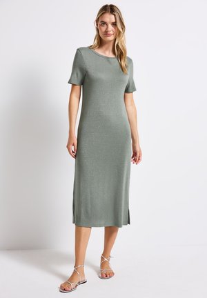 Strickkleid mit kurzen Ärmeln in gedämpftem Grün, knielang mit seitlichen Schlitz; glatte Textur mit dezentem Glanz; kombiniert mit silbernen Riemchensandalen.