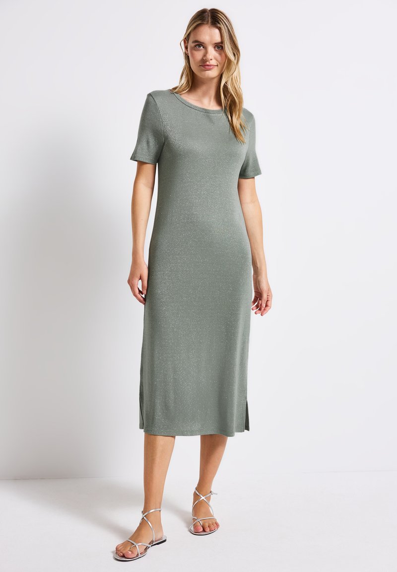 Dzianinowa sukienka z krótkim rękawem w stonowanej zieleni, do kolan, z bocznymi rozcięciami; gładka faktura z delikatnym połyskiem; połączona ze srebrnymi sandałami na paski.