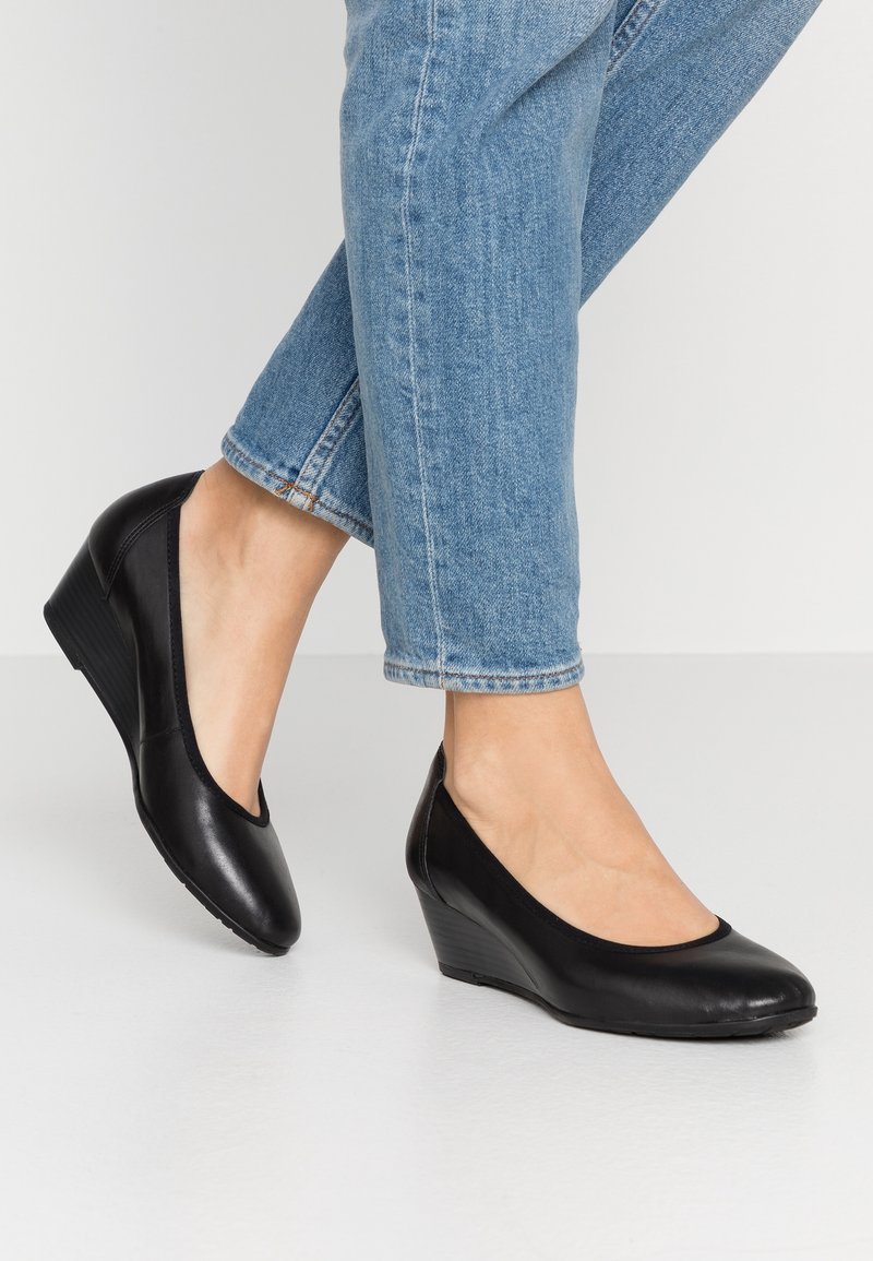 Chaussures à talons compensés en cuir noir avec un bout arrondi et une texture lisse. Accentuées par un talon compensé bas et un design minimaliste.