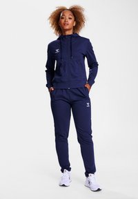 Het marineblauwe trainingspak heeft een rits-houthi met voorzakken en een logo, gecombineerd met bijpassende broek. De stof is glad en het ontwerp is aansluitend.