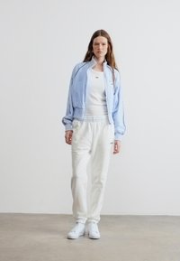 Γυναίκα που στέκεται φορώντας ένα ανοιχτό γαλάζιο μπουφάν με φερμουάρ, λευκή μπλούζα χωρίς μανίκια, λευκά αθλητικά παντελόνια και λευκά αθλητικά παπούτσια μπροστά σε ένα απλό λευκό τοίχο.