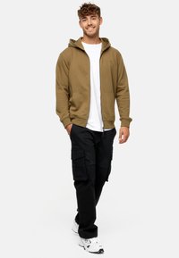 Oliven grøn zip-up hoodie med en frontlomme, parret med sorte cargo bukser. Hvid crew-neck t-shirt under, bæres med hvide sneakers.