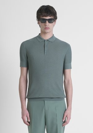 Groene geribbelde polo met korte mouwen, voorzien van een kraag en knoopsluiting. Gecombineerd met bijpassende groene broek. Zonnebril op.