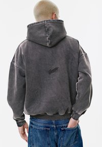 Hanorac gri, supradimensionat, cu glugă, dintr-un material moale și cu manșete ribbate, având un mic logo brodat negru pe spate. Blugi din denim sunt vizibili.