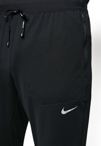 Zwarte sportbroek gemaakt van gestructureerde stof, met een elastische tailleband met trekkoord en een ritszak aan de zijkant. Maat logo op de dij.