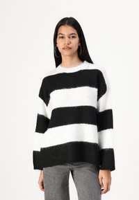 Black and white striped sweater with a loose fit, made of fuzzy material, featuring a round neck and long sleeves. Worn with grey jeans.