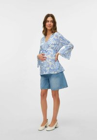 Blusa azzurro chiaro con motivo a foglie, maniche a campana e cintura in vita, abbinata a pantaloni corti di jeans e scarpe con tacco bianco.