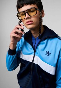 Blauwe en navy tracksuitjas met witte streep; model draagt oversized gele glazen met een ketting; textiel met structuur en logo zichtbaar.