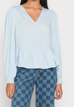 Top peplum azzurro chiaro con maniche lunghe a palloncino, scollatura a V e vita arricciata. Abbinato a jeans a vita alta a quadretti blu.