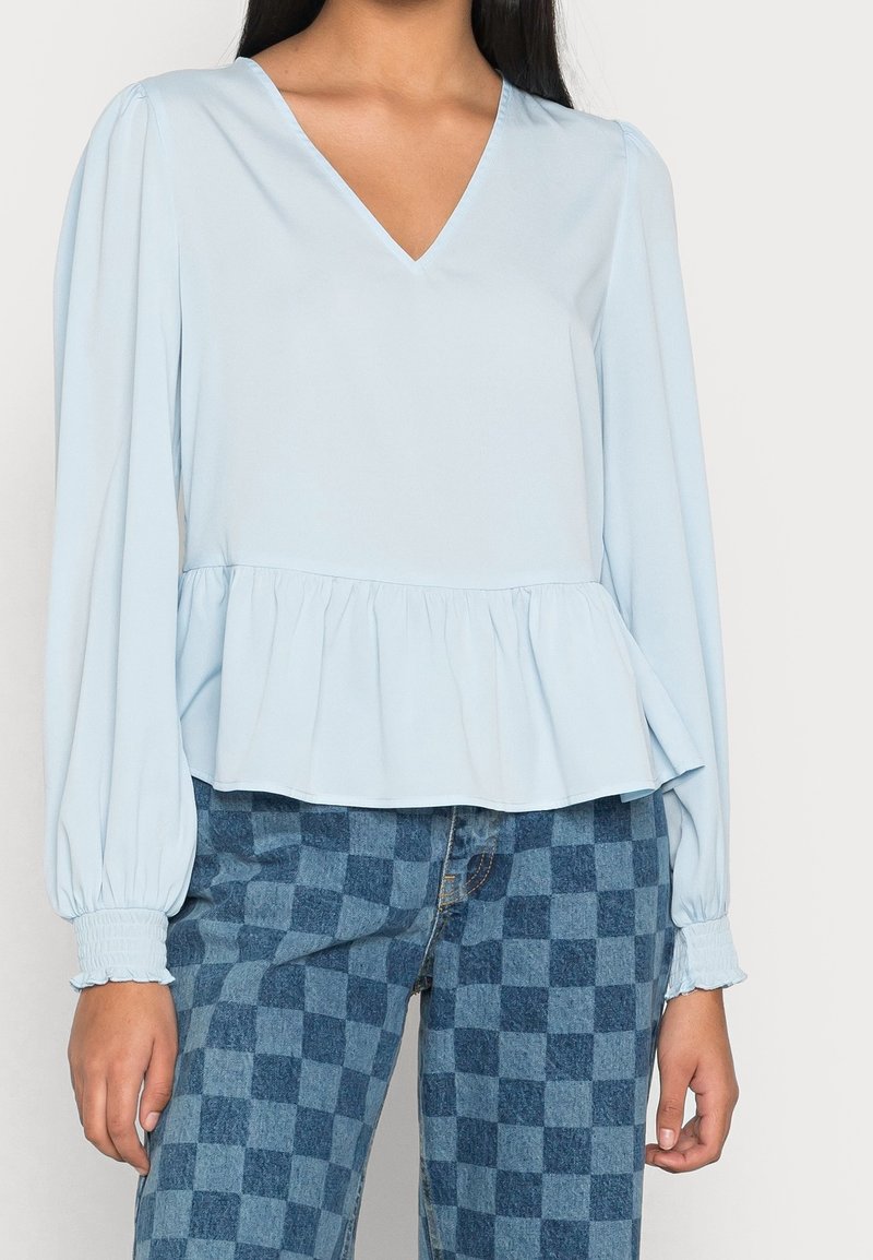 Light blue peplum top with long balloon sleeves, V-neckline, and gathered waist. Paired with blue checkered high-waisted jeans.