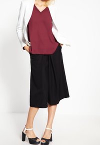 Haut bordeaux à col en V associé à un pantalon culotte noir à jambes larges et une veste blanche. Sandales à plateforme noires à talon épais et brides à la cheville complètent le look.