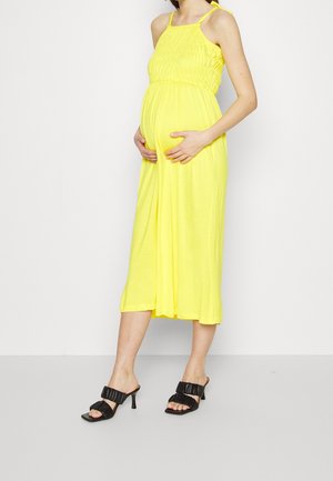 Schwangere Frau in einem gelben, ärmellosen Midikleid, die ihren Bauch hält, trägt schwarze Sandalen mit Absatz vor einem einfarbigen Hintergrund.