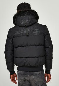 Chaqueta acolchada negra con capucha forrada de pelo sintético, que presenta un sutil patrón de camuflaje en los hombros y puños acanalados.