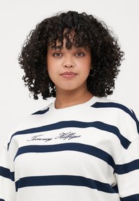 Hvit hettegenser med marineblå horisontale striper, med "Tommy Hilfiger" brodert i skrifttype på brystet. Myk, teksturert stoff.