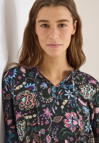 Jeune femme aux longs cheveux bruns portant un haut noir à motifs floraux, debout contre un mur blanc, regardant la caméra.