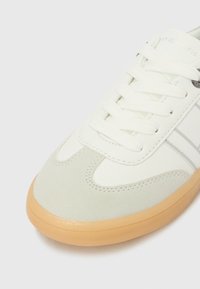 Άσπρο και ανοιχτό γκρι αθλητικό παπούτσι με υφή στη σόλα και κορδόνια, με λεπτές ραφές και μεταλλικό μάτι με το λογότυπο στο πλάι.