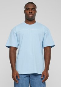 T-shirt surdimensionné bleu clair en coton, avec des manches courtes et un col rond, portant un logo en script blanc sur le devant.