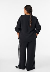 Žena s dlhým vrkočom v čiernom voľnom teplákovom oblečení s bielymi pruhmi, stojaca s rukami v vreckách na bielom jednoduchom pozadí.