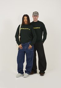 Grüne Sweatshirts mit gelbem Logo, blaue lockere Jeans, graue Mütze, hellgraue Slipper. Einfaches Design auf neutralem Hintergrund.