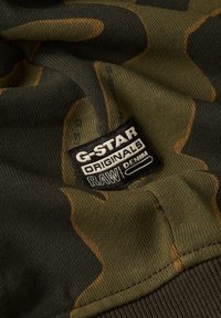 Tela con un patrón de camuflaje en verde oliva con matices de marrón. Presenta una etiqueta negra con texto blanco que dice "G-Star RAW DENIM."