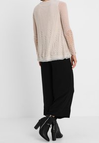 Ljus beige stickad cardigan med intrikata spetsdetaljer vid fållen och ärmarna, ihopparad med lösa svarta byxor och svarta klackade ankelstövlar.