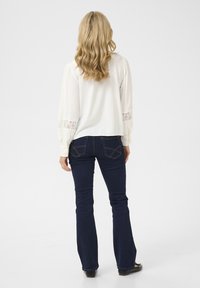 Blouse blanche à manches longues avec des accents en dentelle aux poignets, associée à un jean évasé bleu foncé, vue de dos. Texture lisse, coupe décontractée.