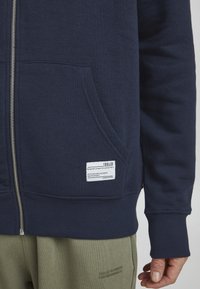 Morska zip-up dukserica napravljena od mekanog materijala, s kengur džepom, rebrastim rukavima i etiketom na lijevom džepu sa uputama za njegu.