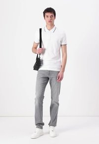 Balta polo marškinėliai su apykakle, apjuostais juodu kraštu, derinami su šviesiai pilkais džinsais ir baltais sportbačiais. Modelis laiko juodą per petį nešiojamą maišą.