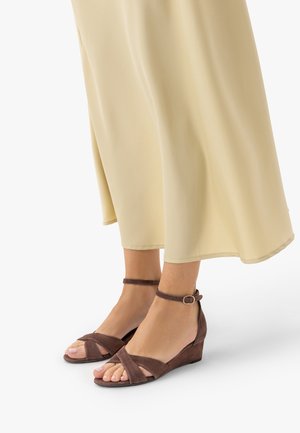 Stopy w brązowych sandałach na koturnie z paskami wokół kostki, widoczne pod płynącą beżową spódnicą lub sukienką.