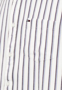 Λευκό πουκάμισο με κουμπιά και κάθετες μπλε και μαύρες ρίγες, τσέπη στο στήθος και ένα μικρό ορθογώνιο λογότυπο σε κόκκινο, λευκό και μαύρο χρώμα πάνω από την τσέπη.