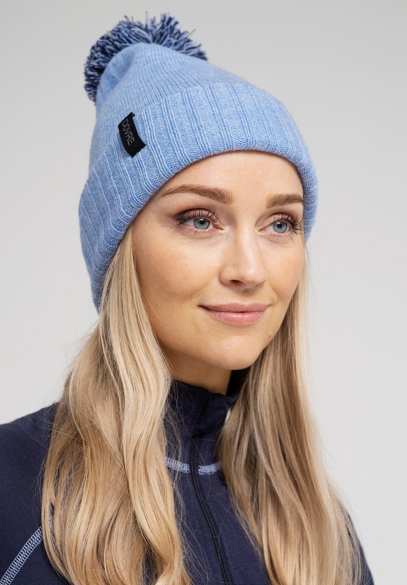 Blå strikket hue med ribbet tekstur og en topp pompom, der har et lille sort mærke på siden. Velegnet til koldt vejr.