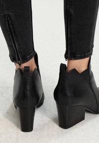 Botas de cuero negro hasta el tobillo con un tacón más grueso, puntera afilada y detalle de cremallera en la parte trasera, que exhiben un acabado suave y un diseño estructurado.