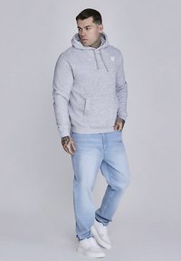 Lichtgrijze hoodie met een buidelzak en trekkoord. Gecombineerd met lichtblauwe spijkerbroek en witte sneakers. Soepele katoenen textuur.