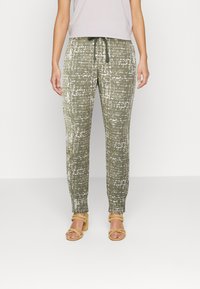 Calças texturizadas em verde-azeitona com um padrão abstrato branco, com cintura elástica e cordão ajustável. Combinadas com sandálias bege claras.
