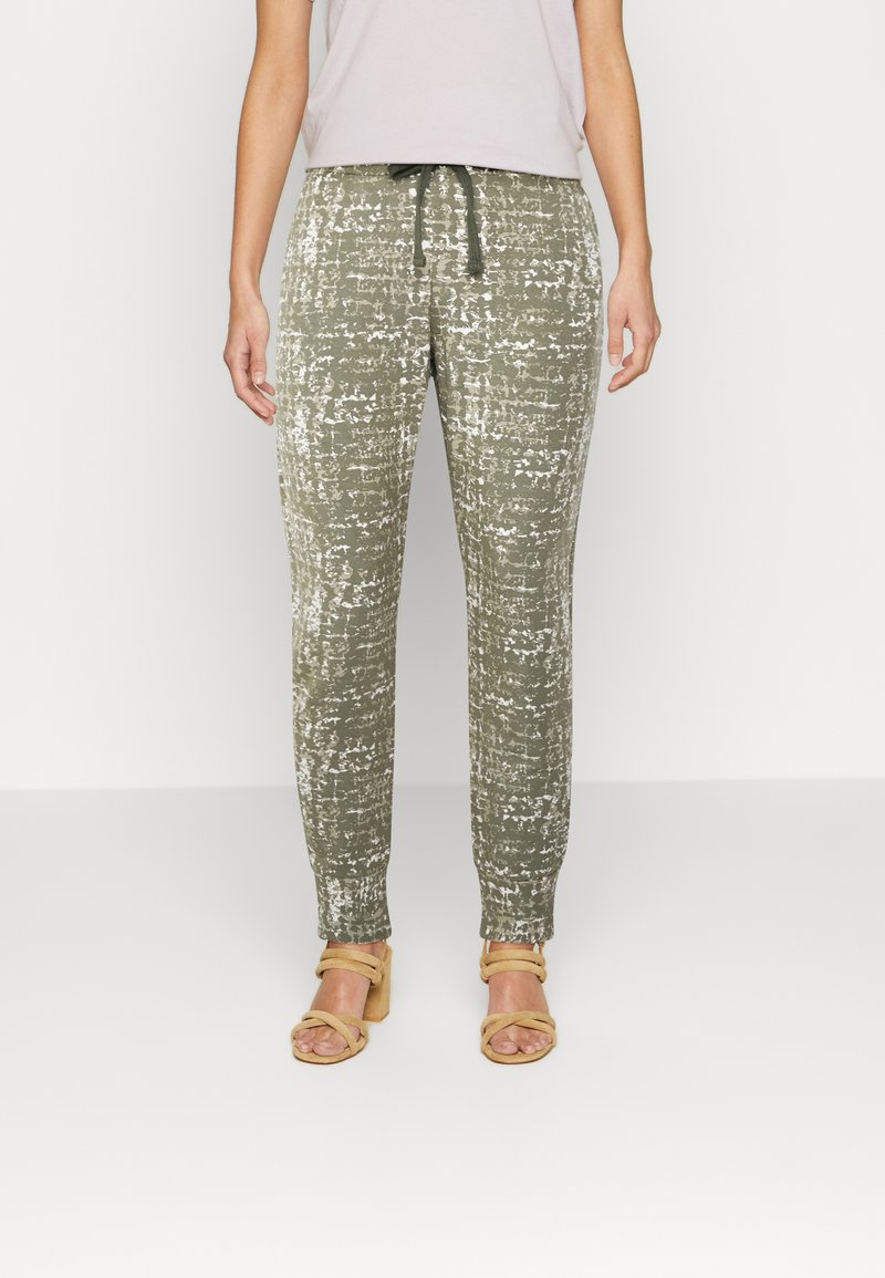 Calças texturizadas em verde-azeitona com um padrão abstrato branco, com cintura elástica e cordão ajustável. Combinadas com sandálias bege claras.