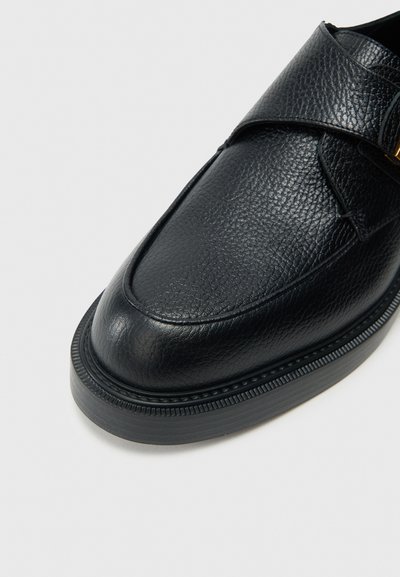 Čierny kožený mokasín s textúrovaným povrchom, zaoblenou špičkou a nastaviteľným remienkom so zlatým zdobením. Hrubá, textúrovaná podrážka.