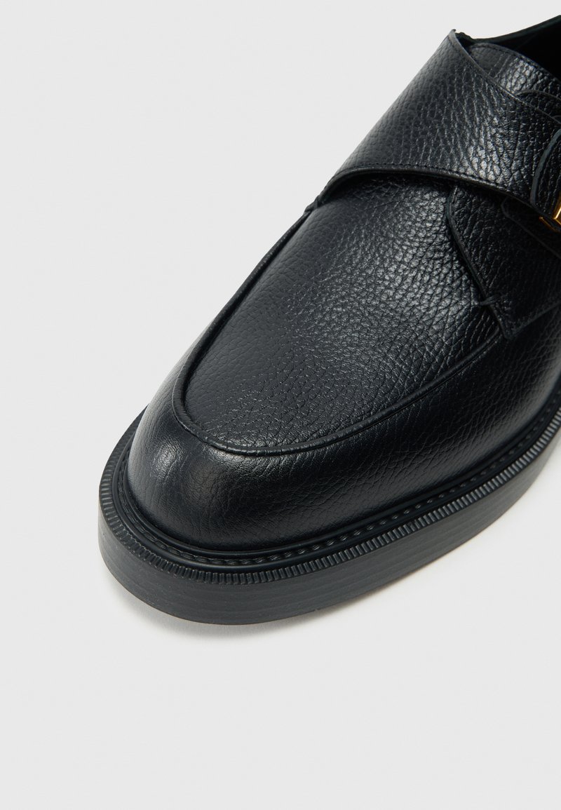 Mocassino in pelle nera con finitura strutturata, punta arrotondata e cinturino regolabile con dettaglio in metallo dorato. Suola spessa e testurizzata.