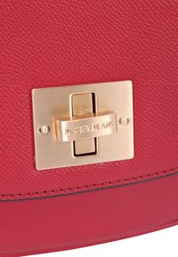 Sac en cuir texturé rouge doté d'un fermoir rectangulaire doré avec "JOOP! JEANS" gravé. Des détails de couture noirs ajoutent une définition supplémentaire.