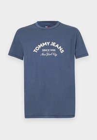 Mėlynas trumpas rankovėmis marškinėliai su baltais užrašu "TOMMY JEANS Since 1985 New York City" išspausdintu viduryje priekyje.