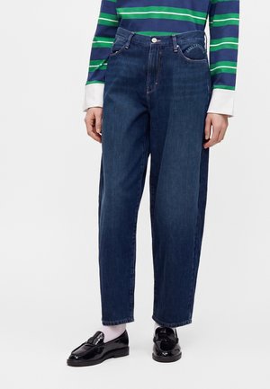 Osoba nosząca ciemnoniebieskie dżinsy denim, długi top w zielono-niebieskie paski z białymi mankietami, białe skarpetki i czarne błyszczące mokasyny.