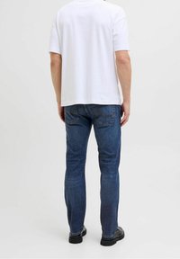 Camiseta blanca de algodón con mangas cortas, combinada con jeans de mezclilla azul que tienen bolsillos traseros y detalles de costuras. Se ven zapatos negros.