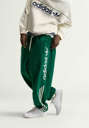 Persona que lleva pantalones deportivos verdes de Adidas con texto y rayas blancas, una sudadera cremosa de Adidas y zapatillas blancas sobre un fondo liso.