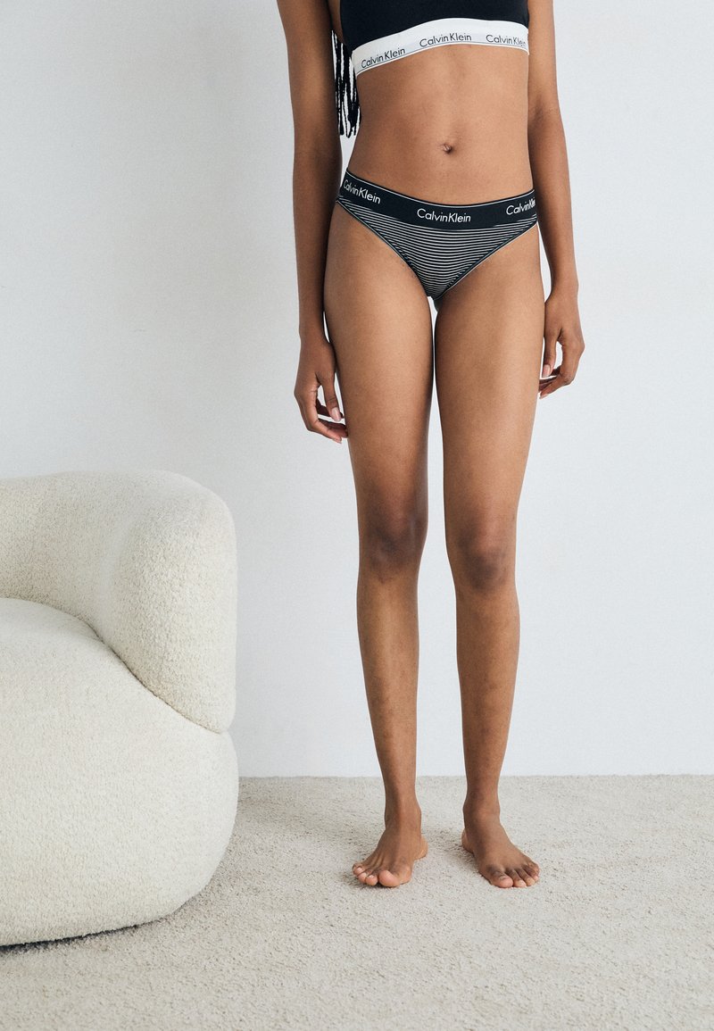 Calvin Klein black and white striped thong with logo waistband, paired with a black sports bra. Smooth fabric, minimalistic design, and no patterns.