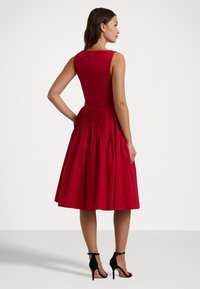 Rochie roșie fără mâneci, cu un trunchi ajustat și fustă pliată. Închidere cu fermoar la spate, lungime până la genunchi, asortată cu pantofi cu toc negri. Textură fină.