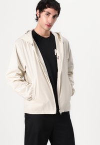 Light beige, zip-up jacket with a hood, crafted from a smooth, shiny material. Features side pockets and a minimalist design.