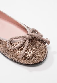 Bailarinas con un exterior de lentejuelas bronce, ribetes grises y un detalle de lazo de tela en la puntera. Forro interior suave en color rosa y suela negra.